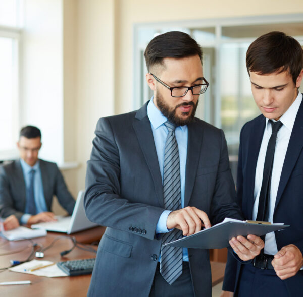 Confident businessman pointing at document while explaining his idea to his partner on background of their colleagues