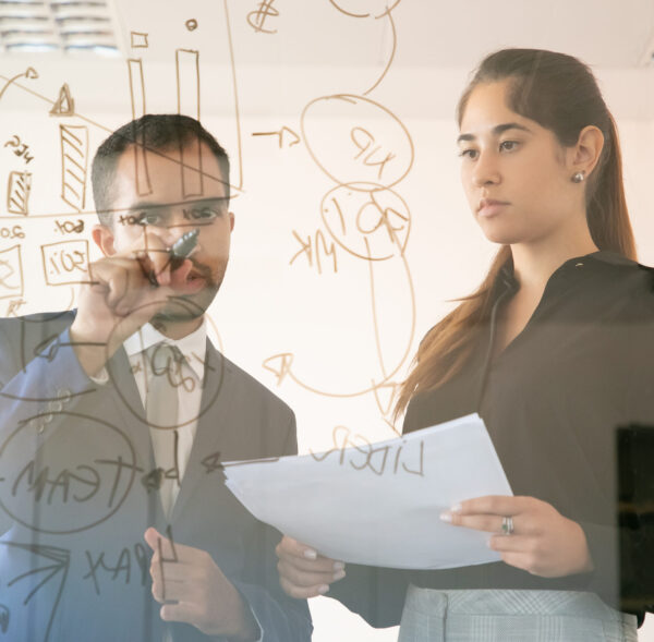 Content African American manager writing chart on glass board. Professional young pretty female colleague holding document and looking at graph in conference room. Teamwork and marketing concept